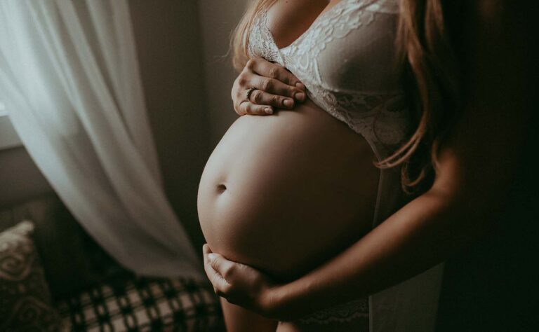 mom-to-be holds big belly near the window at home during the matenrity photo session with victoria vasilyeva