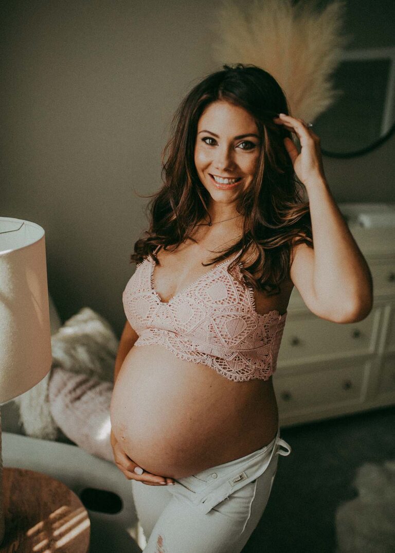 mom-to-be with long dark hair in light pink crochet top and blue jeans smiles to the camera. The portrait of pregnant mama was taken near prenatal yoga raleigh nc.