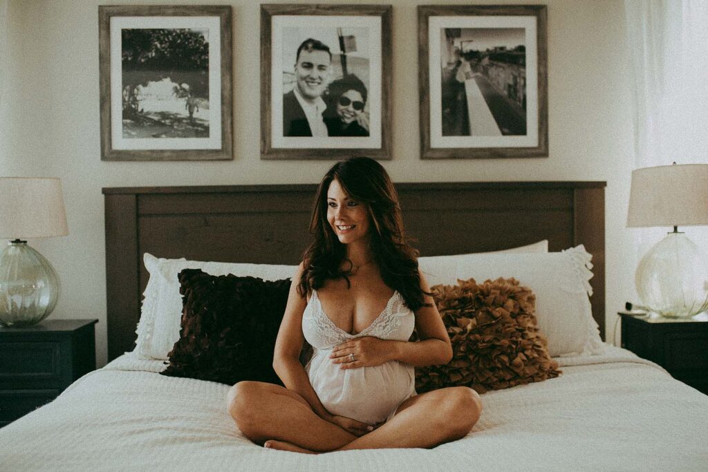 mom-to-be in lace white robe is sitting on a bed and holding her bump