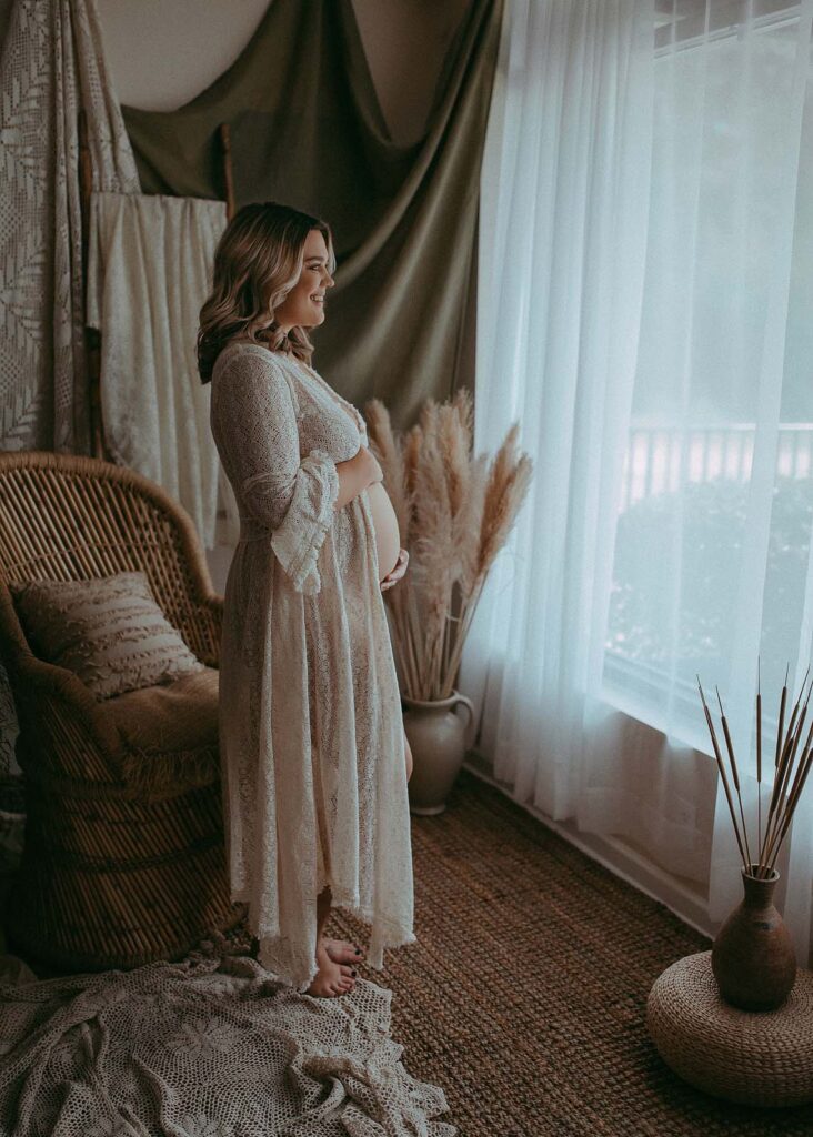 mom-to-be in wtite lace robe is standing near the window in photo graphy studio in Apex