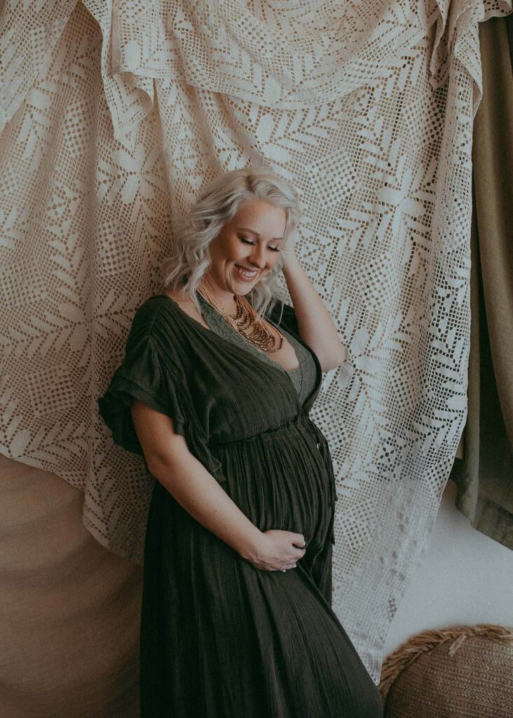 Pregnant woman stays near the wall, holds her belly, and giggle down. Maternity portrait session took place at Victoria Vasilyeva Photography studio near prenatal massage Durham NC