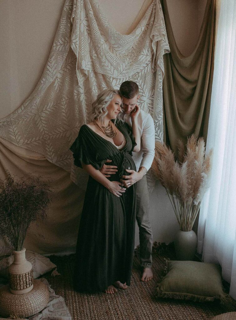 Expecting couple is standing near the window, hugging each other and holding big pregnant belly during maternity photo session with Victoria Vasilyeva Photography.