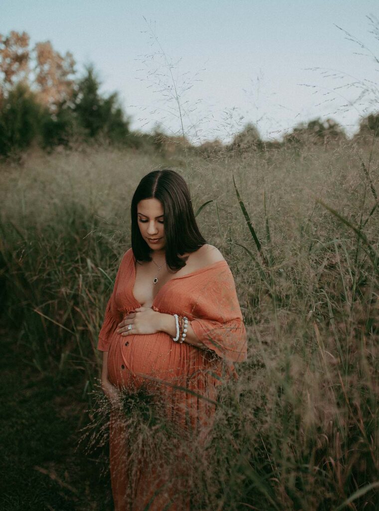 Pregnant women in orange boho dress with black hair is among high grass looking down the belly, holding it. Maternity session took place near prenatal massage Raleigh NC studio.