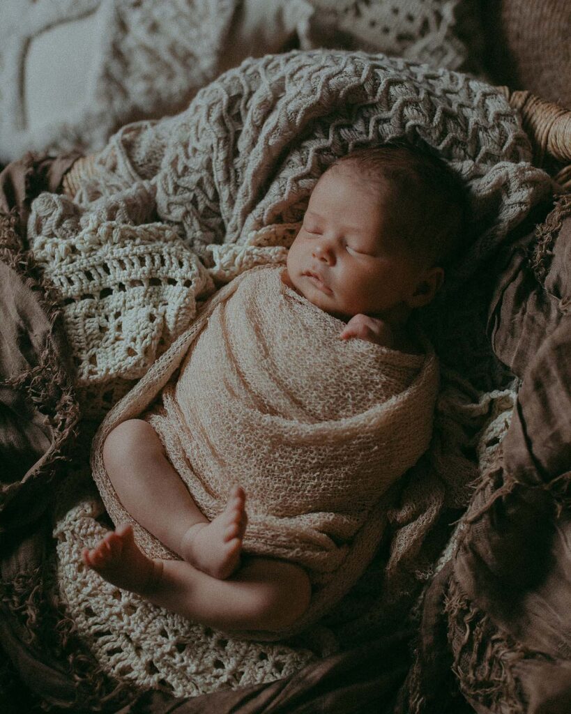 9 days old baby wrapped in tan knitted blanket lays on rattan chair in Apex Photo Studio.