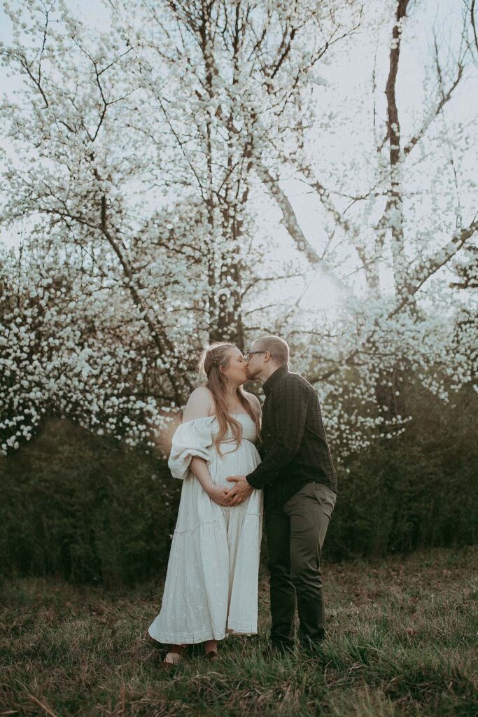 Happy expecting parents are kissinf under huge blooming tree at NCMA in Raleigh. Maternity photo session by Victoria Vasilyeva Photography.