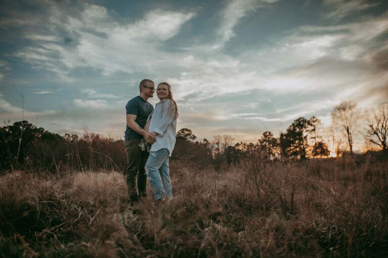 A photograph of a pregnant couple standing in front of each other was made by Raleigh maternity photographer - Victoria Vasilyeva Photography. The pregnant mama is wearing a white blouse and blue jeans, while the man is dressed in a blue t-shirt and dark green pants. The couple is standing in the middle of a huge field at NCMA with a gorgeous sunset in the backdrop. The expecting parents hold white flowers and have their hands resting under the belly.