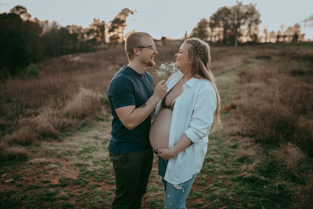 A photograph of a pregnant couple standing face to face and smiling at each other was made by Raleigh maternity photographer - Victoria Vasilyeva Photography. The woman is visibly pregnant, wearing a white blouse and blue jeans, while the man is dressed in a blue shirt and dark green pants. The couple is standing in the middle of a huge field at NCMA. The woman has her hand resting under her belly, suggesting a sense of pride and anticipation for the new arrival.