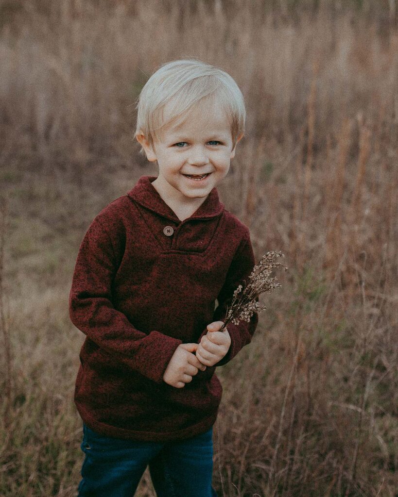 Portrait of a blond-haired and blue-eyed boy 4 years old. He collected a beautiful bouquet of wild flowers for his mother. He is very happy and smiles at the camera. The photo was taken during the family photo session at Anderson Point Park in Raleigh, NC by Victoria Vasilyeva Photography.
