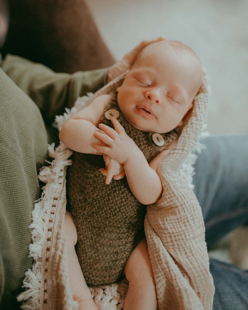 Dad holds his newborn son in a green romper in his arms. The baby fell asleep. He sleeps soundly with his arms folded across his chest. The portrait was taken by the best newborn photographer - Victoria Vasilyeva Photography.