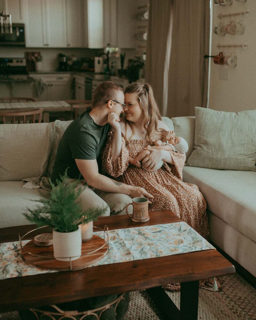 Young parents are sitting on the sofa in the living room. Mom in a beautiful long dress holds her daughter in her left hand and strokes her husband with her right hand. They are in love and happy. The portrait was taken by the best baby photographer - Victoria Vasilyeva Photography.
