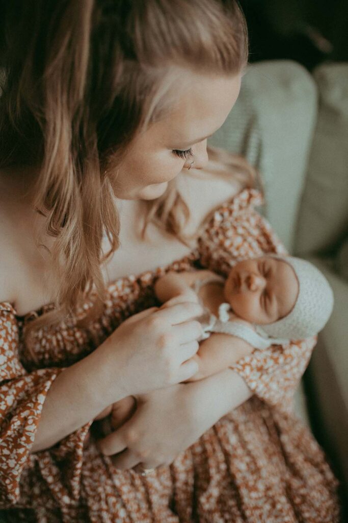 The young beautiful mother with long blond hair and black eyelashes tenderly looks at her newborn daughter. The baby sleeps sweetly in her arms. The portrait was taken by the best newborn photographer - Victoria Vasilyeva Photography.