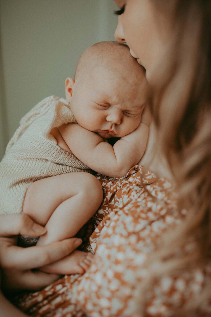 A baby with plump cheeks in a knitted light romper sleeps on her mother's shoulder. The mother turned her head and kissed her daughter on the forehead. Photo taken by the best newborn photographer - Victoria Vasilyeva Photography near Triangle Physicians for Women.