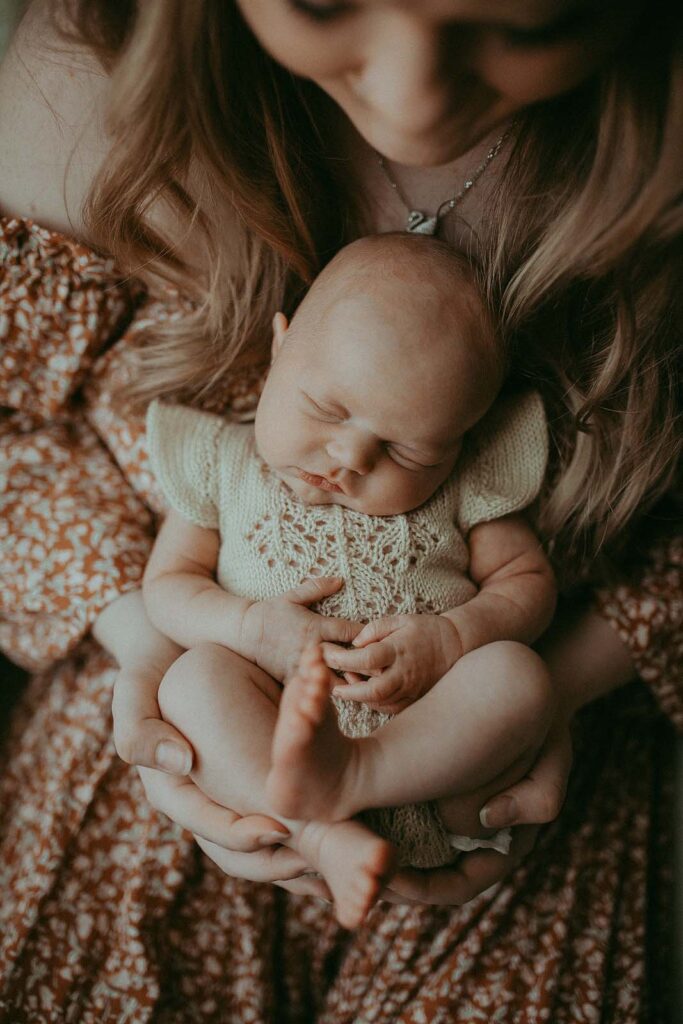 A baby in a knitted light romper, cross-legged, sleeps in her mother's arms. Photo taken by the best newborn photographer - Victoria Vasilyeva Photography.