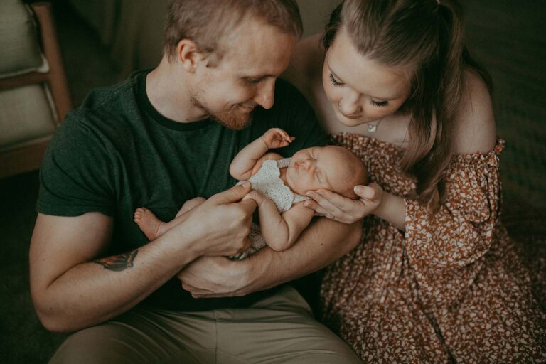 Mom and dad are holding their newborn daughter in their arms. The baby sleeps sweetly, and the parents look down at her and admire her. Photo taken by the best newborn photographer - Victoria Vasilyeva Photography.