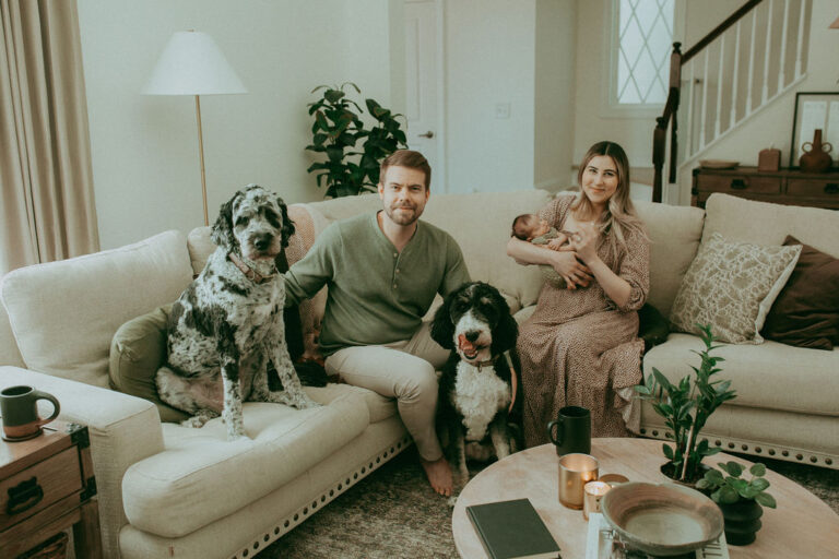 Family portrait of mom, dad, newborn, and 2 curly dogs. The family sits on a couch in their living room. The baby boy was born just some days ago. The family prepared for his birth by shopping at Precious Arrow.