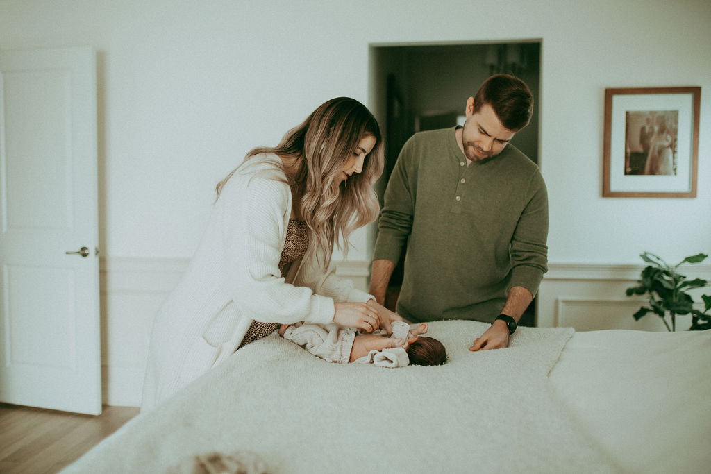 Mom with long beautiful hair is changing the diaper of her newborn baby boy. Dad in muted green hanley is staying next to her and looking at the baby. This family portrait was taken during newborn photo session with Victoria Vasilyeva Photography