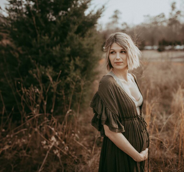 A pregnant woman with short blonde hair stands gracefully in a boho maxi dress, surrounded by tall grass, near the Blissful Births in Raleigh. The serene setting encapsulates the anticipation and beauty of the maternity journey.