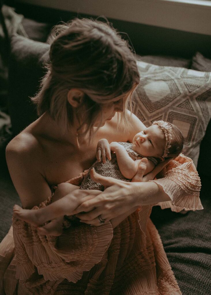 A photo of a mother with short curly blonde hair cradling her newborn baby girl near a sunlit window after their visit to Chapel Hill Midwives.