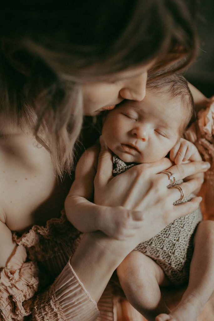 An image featuring a mom with adorable short curly blonde hair, holding her precious newborn baby girl, basking in the soft natural light by a window following their visit to Chapel Hill Midwives.
