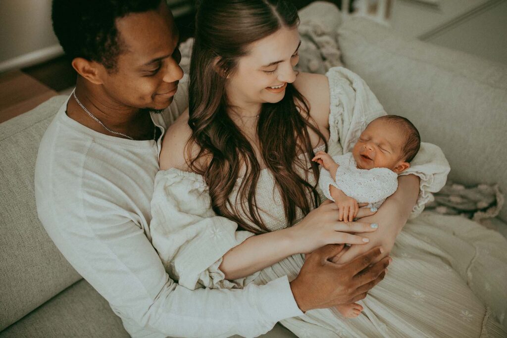 First photo session of 4 days old boy with his mom and dad. They are sitting on a couch in their living room. Mom with long beautiful hair holds baby and looks at him.