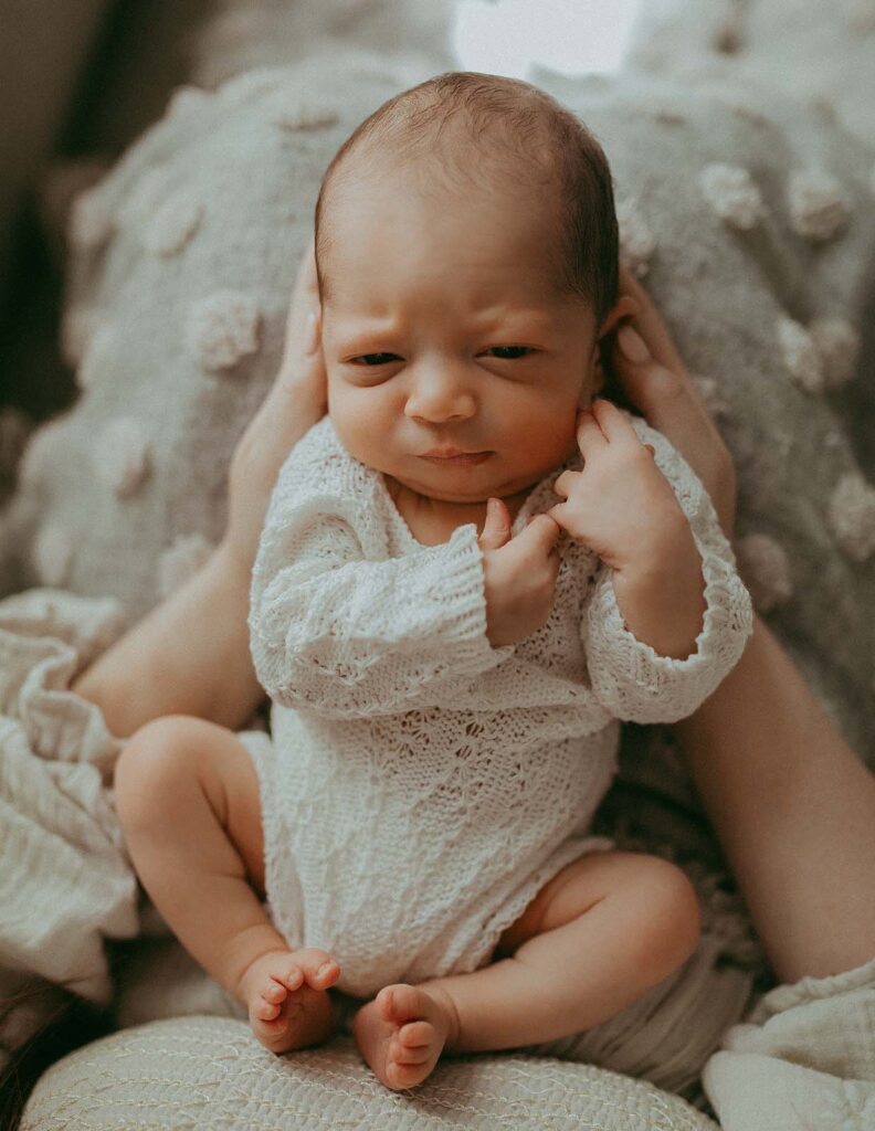 A portrait of a newborn boy. He is dressed in a white romper. The baby is lying on his mother's lap. Mom hugs him with both hands. Newborn portrait was taken by Victoria Vasilyeva Photography - Cary newborn photographer.