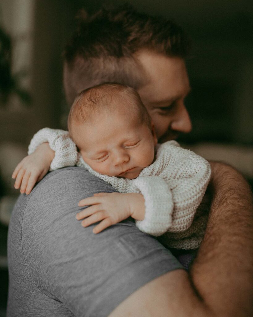 Dad in a gray T-shirt is holding his sleeping son on his shoulder. The baby is only 9 days old. He is dressed in a knitted sweater. The little one sweetly sleeps, turning his head to the left. This family portrait was taken during the newborn photo session with Victoria Vasilyeva Photography.