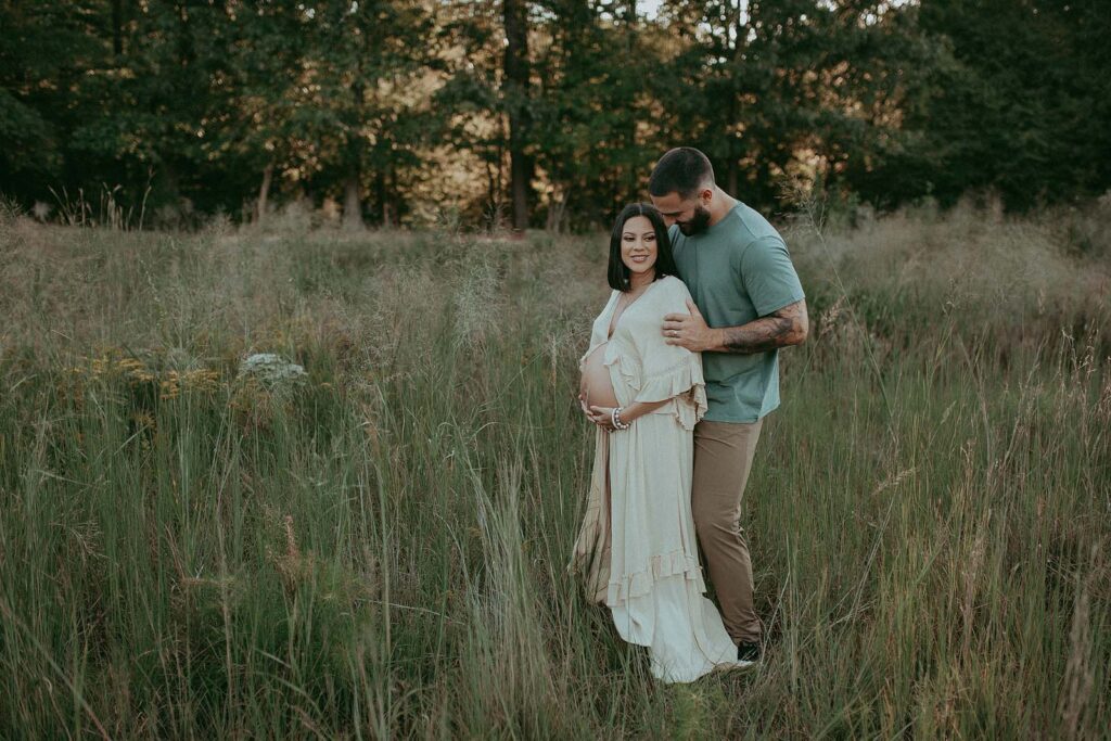 In a picturesque setting of lush green grass, a radiant mom-to-be embraces her pregnancy glow with her husband, showcasing her boho style in a flowing maxi dress, as captured by Victoria Vasilyeva Photography during a maternity photo session.