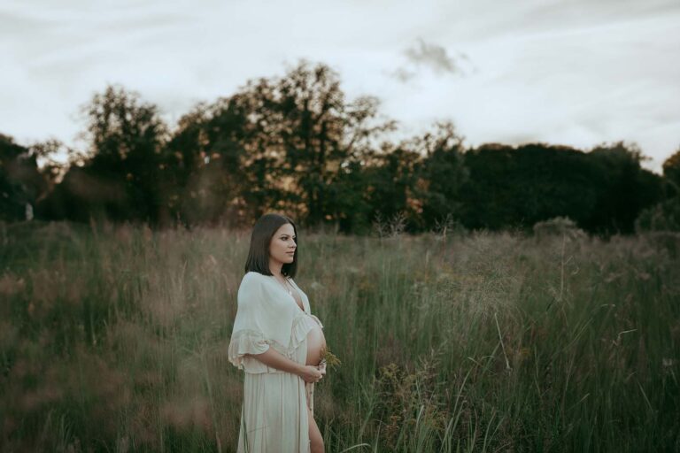 An ethereal image captures a beautiful expectant mother, adorned in a boho maxi dress, standing amidst tall grass during a serene maternity photo session with Victoria Vasilyeva Photography.