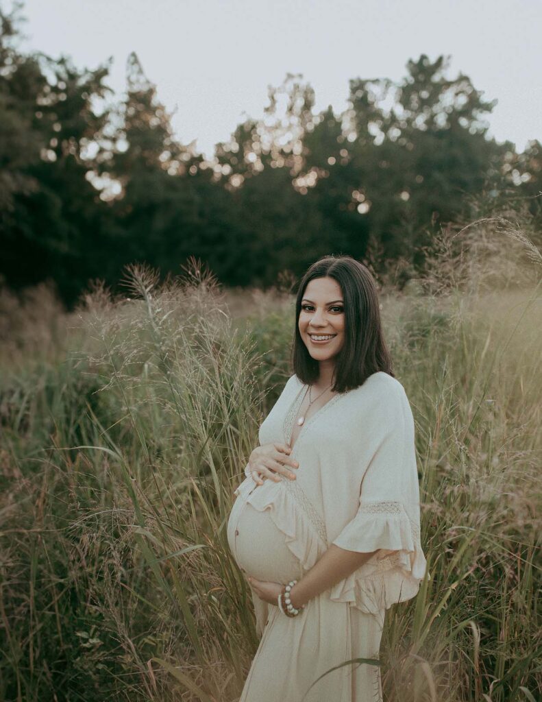 A stunning mom-to-be wearing a boho maxi dress poses gracefully in a field of high grass during her maternity photo session with Victoria Vasilyeva Photography.