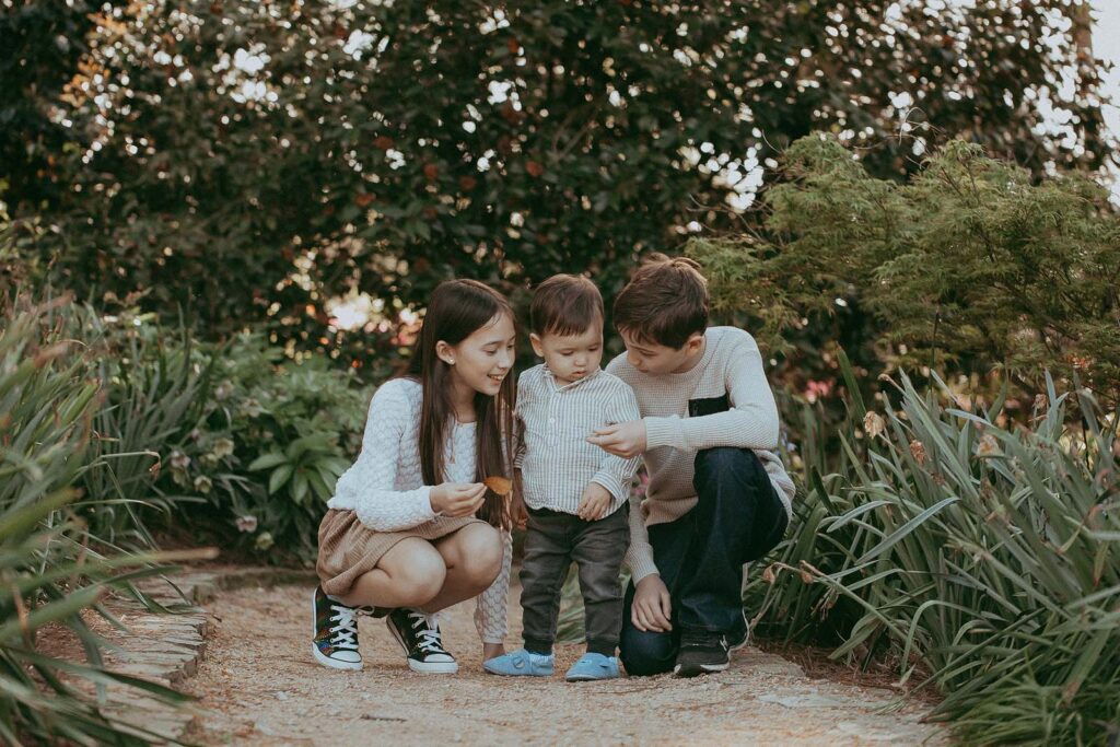 The children are squatting and showing their younger brother their new toy. It was purchased at one of Raleigh toy stores. The photo was taken at WRAL Azalea Garden by Raleigh NC family photographer - Victoria Vasilyeva Photography.