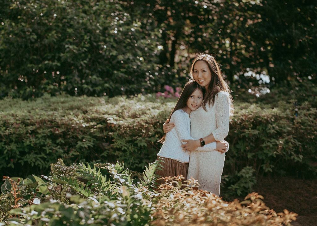 Mom and daughter are posing for Raleigh family photographer - Victoria Vasilyeva Photography. They stand side by side, embracing each other. The girl rests her head on her mother's chest. The setting sun's rays beautifully illuminate their dark and shiny hair from behind.