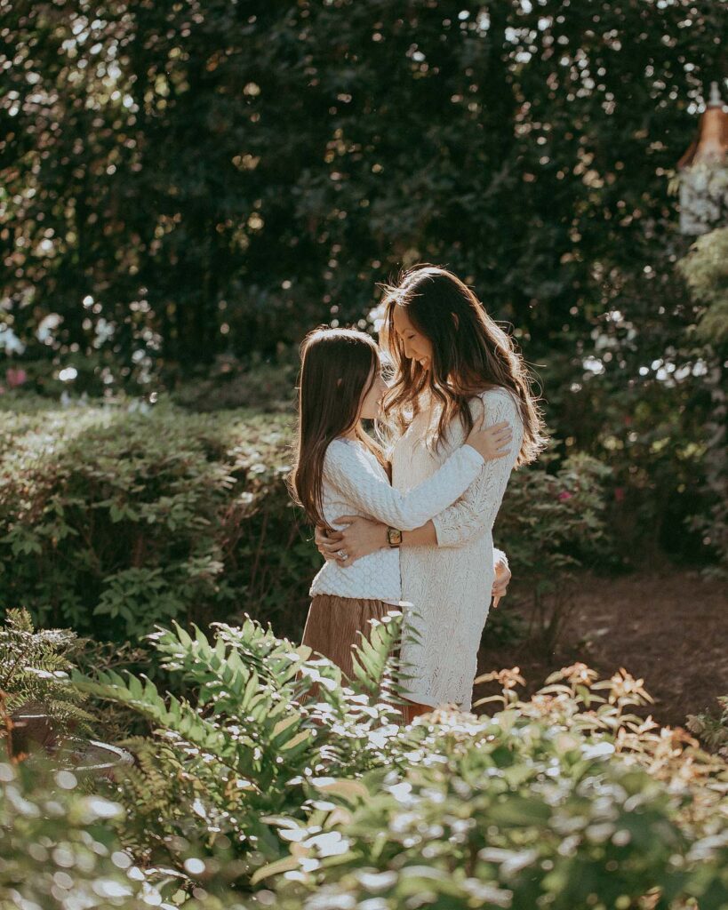 Mom and daughter are posing for Raleigh family photographer - Victoria Vasilyeva Photography. They stand face to face, embracing each other. The girl looks at her mother. The setting sun's rays beautifully illuminate their dark and shiny hair from behind.