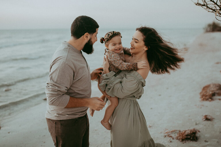 Family enjoying a serene sunset on the beach after a visit to Childtime of Greensboro. Mom, dressed in a stylish sage maxi dress, and dad lovingly hold and embrace their adorable 2-year-old daughter, creating a heartwarming moment of connection and love.