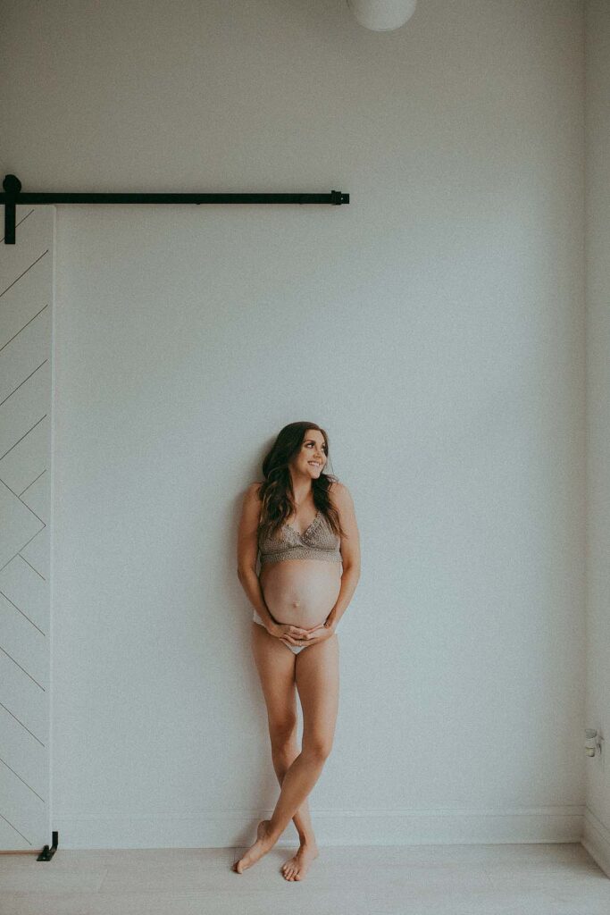 Stunning mom-to-be in boho lace langerie is leaning to the wall and looking to the left at photography studio near Prenatal Yoga Greensboro NC. Maternity photo session by Victoria Vasilyeva Photography.