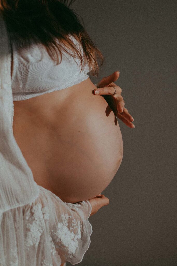 The photo of big belly of pregnant woman in lace white robe. She is holding the belly with her right arm and touching it with point finger. The photo was taken at the photography studio near Prenatal Yoga Greensboro NC by maternity photographer - Victoria Vasilyeva Photography.