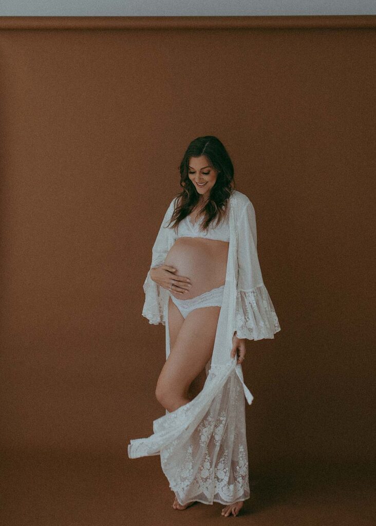 Stunning mom-to-be in white lace robe is dancing , smiling and looking at her big belly. The maternity portrait was taken by Victoria Vasilyeva Photography at Studio 557 in Holly Springs.