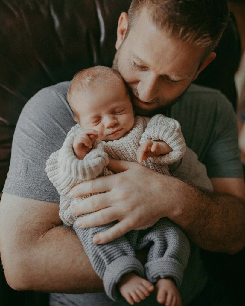 A father and son bonding at home. The father has a beard and wears a grey shirt. The baby boy is wearing a white cozy sweater. Portrait by Fayetteville newborn photographer - Victoria Vasilyeva Photography.