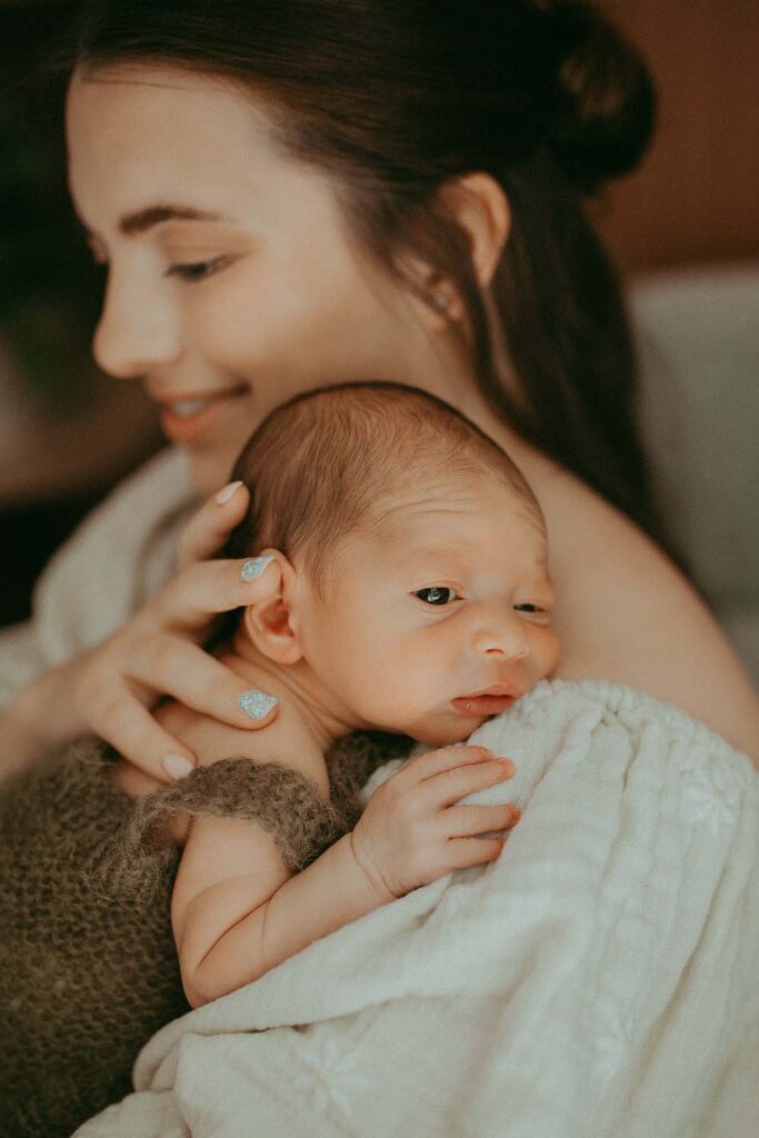 Mommy with long dark hair is holding and hugging her newborn son. He is nealing to her shoulder and looking around. The portrait of mommy and son was taken by Victoria Vasilyeva Photography - Fayetteville Newborn Photographer.