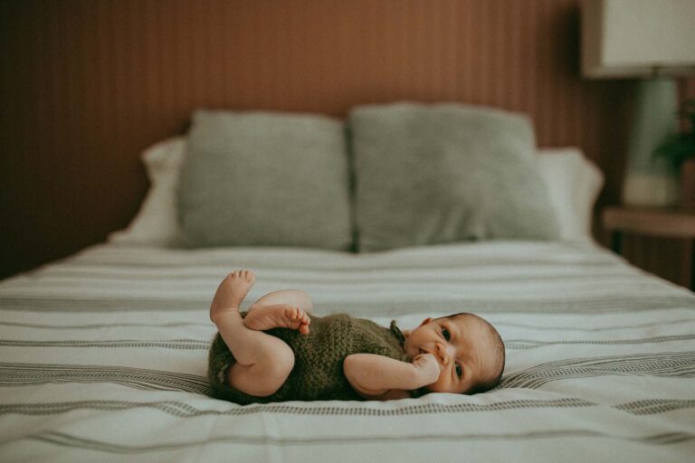 A tiny newborn baby in a knitted green romper is in the master bedroom on a bed. He is wide awake and looking around during his first-ever photo session with Fayetteville Newborn Photographer.