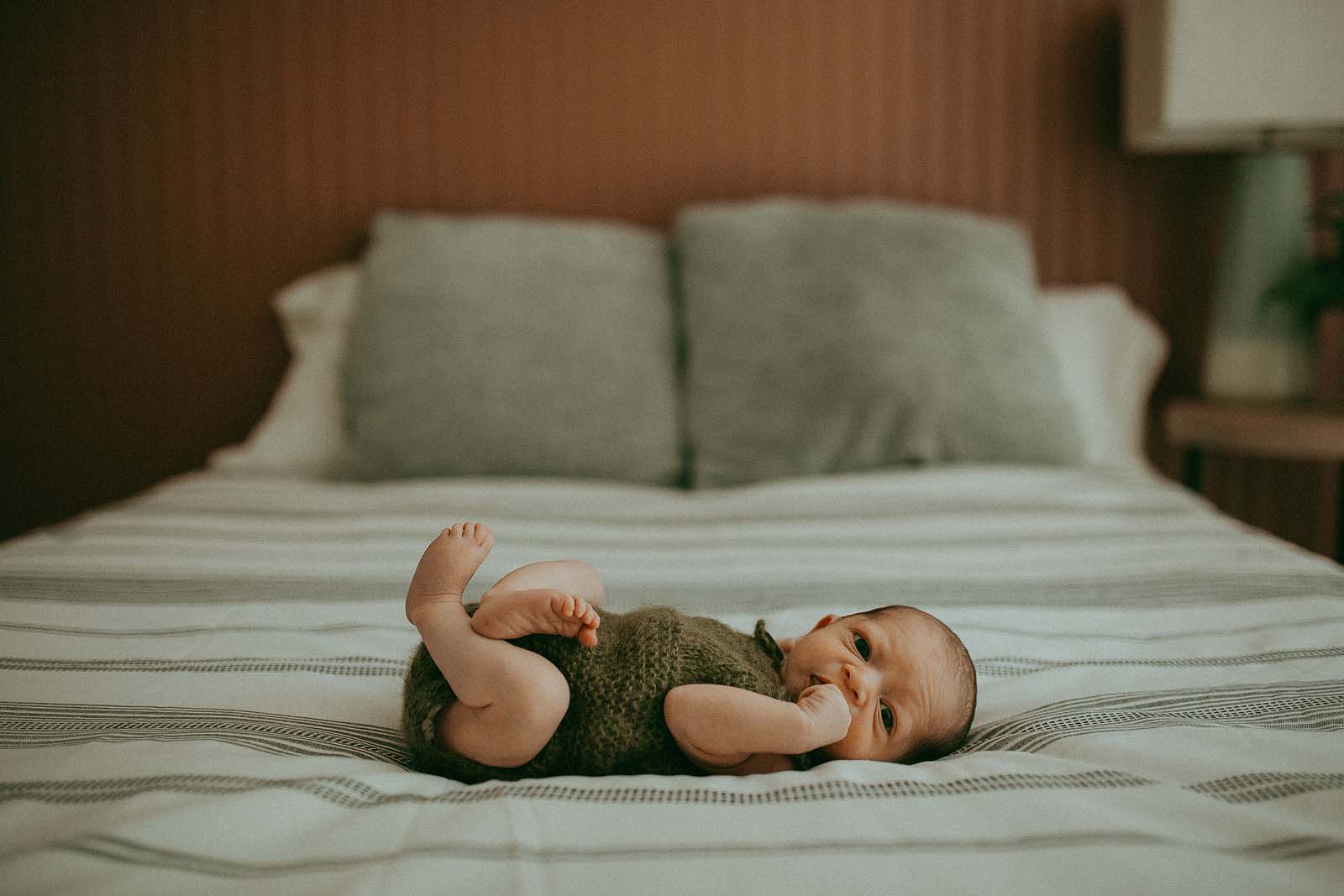 A tiny newborn baby in a knitted green romper is in the master bedroom on a bed. He is wide awake and looking around during his first-ever photo session with Fayetteville Newborn Photographer.