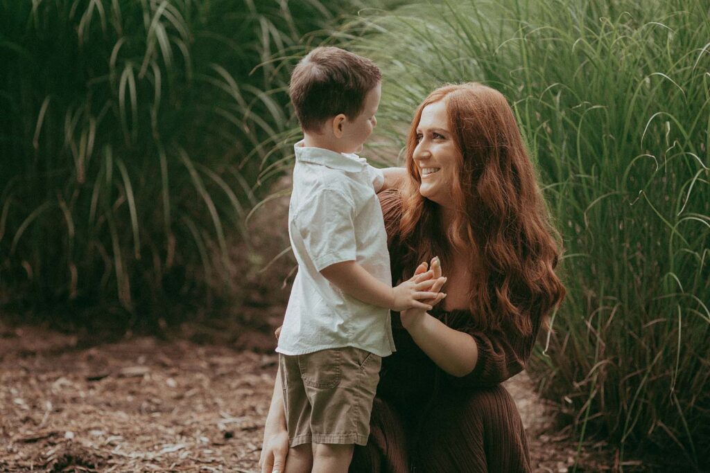 A beautiful mother with long red hair and her son smile to each other. They are standing in front of a high grass. Family portrait by Raleigh family photographer - Victoria Vasilyeva Photography.