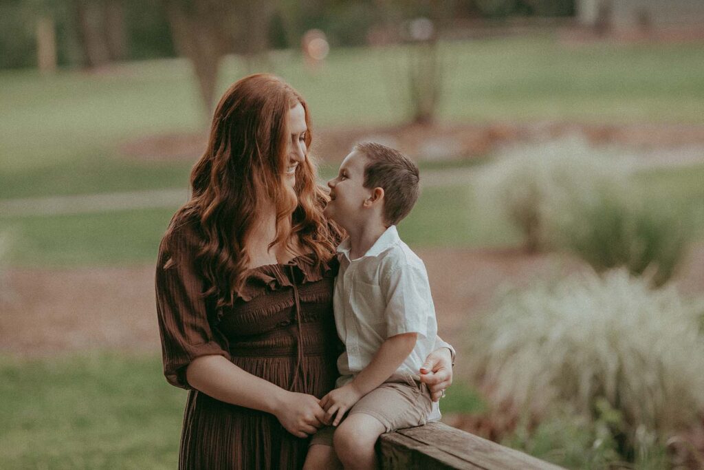 A mother and son pose for a photo near North Raleigh Pediatricians. The mother has long red hair and is wearing a maxi brown dress. The son is wearing a white shirt and tan short.