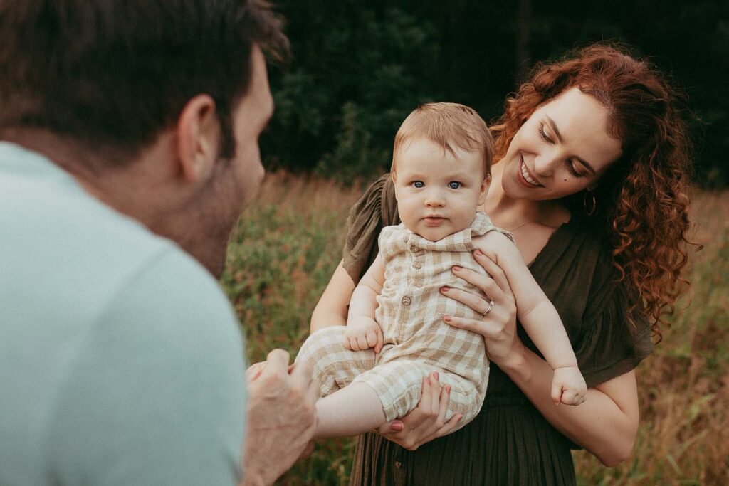 The mother and father hold their baby boy in their arms. The baby in plaid Rylee and Cru romper is looking at the camera. The parents are smiling and looking at him.