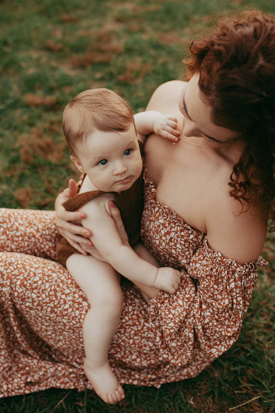 The mother with long red hair and baby play on the grass. The baby is showing his tongue and the mother is smiling. Photo session took place near A Woman's Place in Fayetteville, NC. Photographer - Victoria Vasilyeva Photograthy.