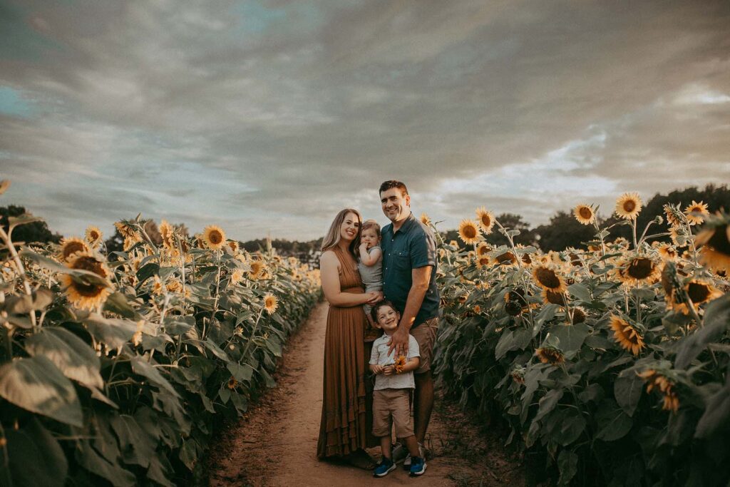 A mother and father hold and hug their boys. The kids are smiling and looking at the camera. The parents are smiling and looking at the camera too. They are standing in a field of sunflowers near indoor playground raleigh nc.