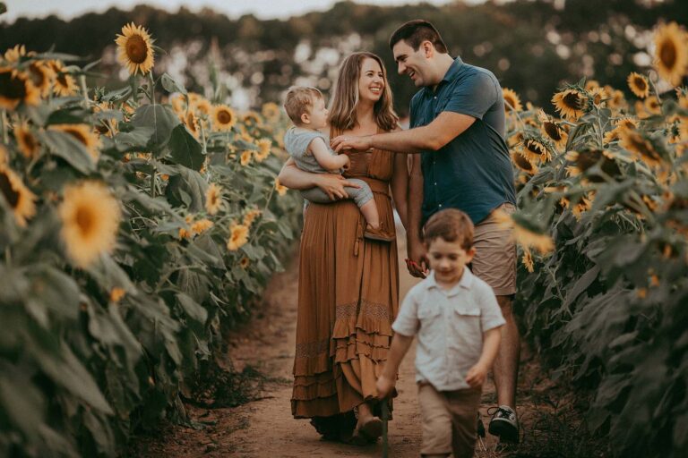 A family of four walks along the sunflower field. The mother is holding the baby in her arms and the father is walking next to them. They are all smiling and looking happy. There is a green forest in the background. Family portrait was taken by Raleigh family photographer near indoor playground raleigh nc.