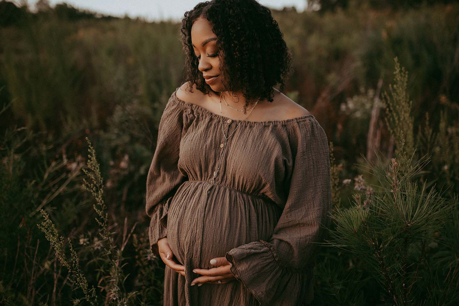 Stunning mom-to-be in boho maxi dress is standing in the field. She is holding her belly with both hands and looking down. She looks very happy. Photo session was taken near Baby Shower Venues Cary NC by Victoria Vasilyeva Photograher.