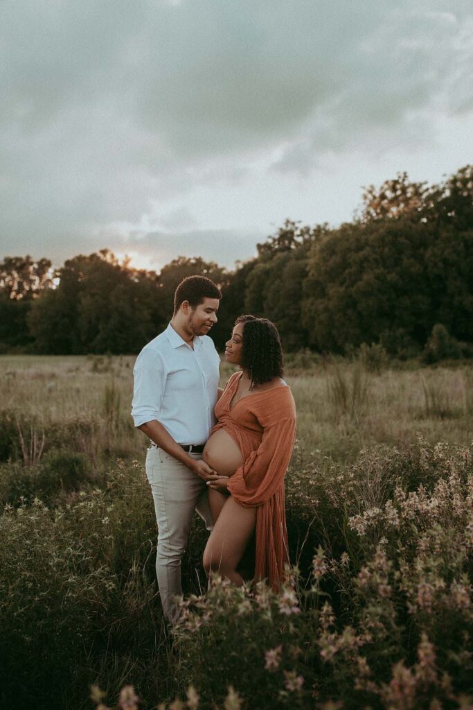 The expecting couple is full of love and happy. They are standing in the field and looking at each other. Mom wears stunning boho maxi dress from Jens Pirate Booty. Maternity photo session wan taken near Baby Shower Venues Durham NC by Victoria Vasilyeva Photography.