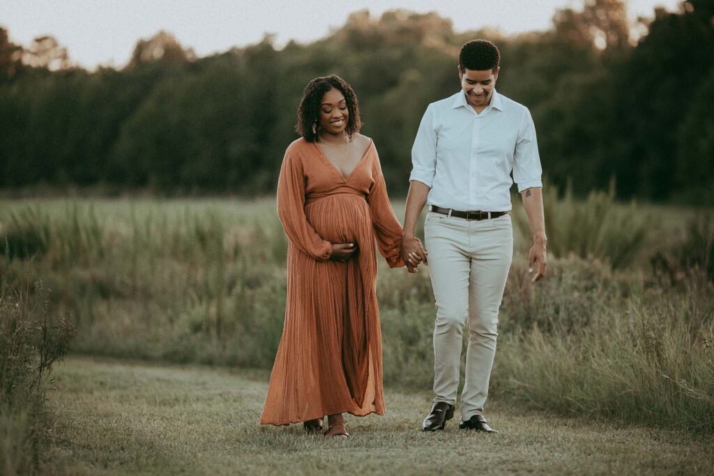 The expecting pearents are holding the hands and walking down the path. They are smiling and looking down. Mom wears boho orange dress from Jens Pirate Booty. Portrait session by Victoria Vasilyeva Photography.