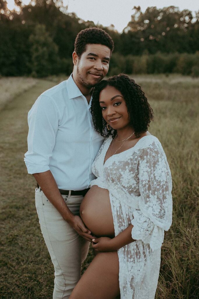 Mom and Dad Glow with Joy in Maternity Photos by Victoria Vasilyeva Photographer. This photo shows a beautiful pregnant mom and her loving husband, who is cradling her belly. They are both wearing white, and the photo is full of love and happiness.